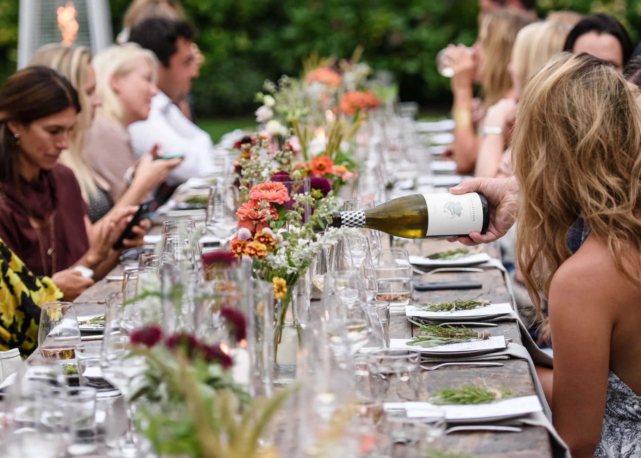 Friends at a long table laid with flowers and wine in a garden